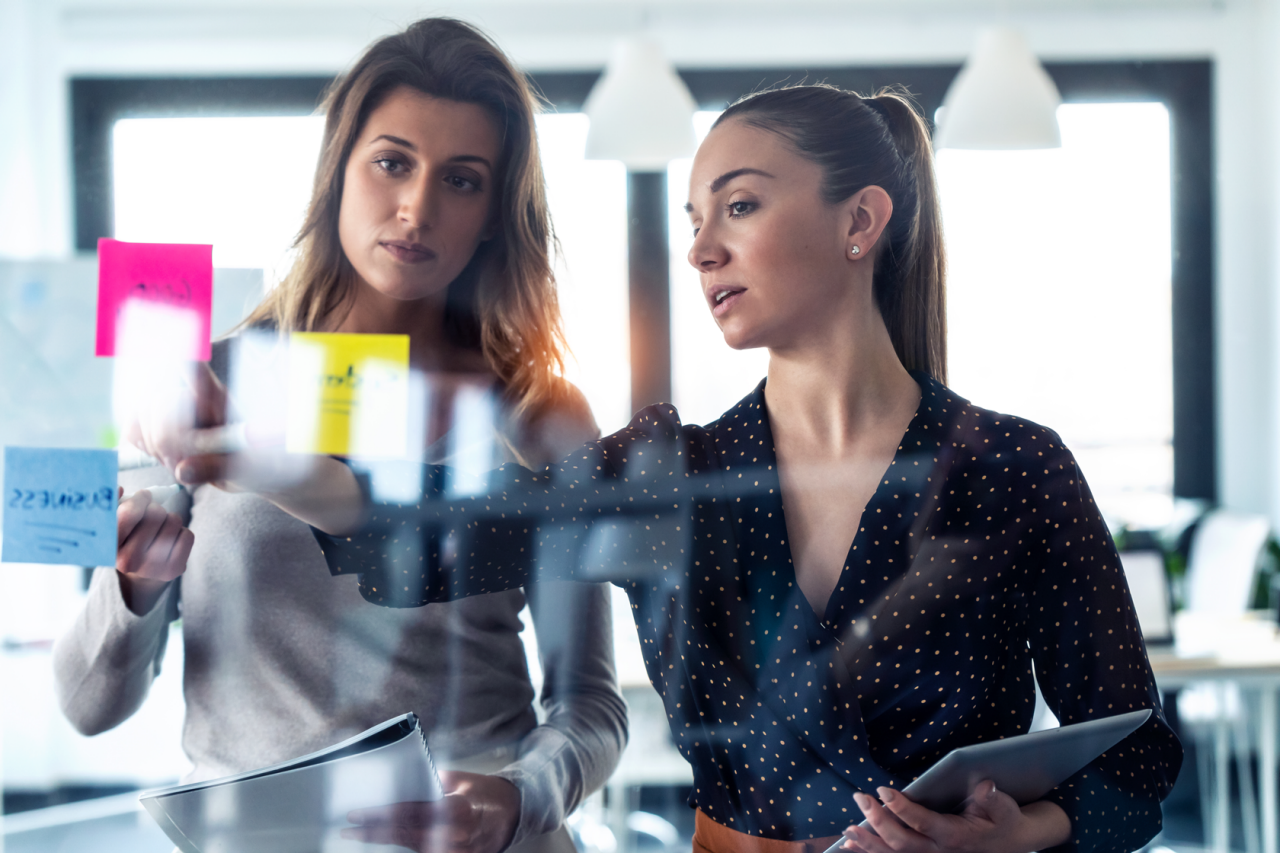 Two business young women working together on wall glass with post it stickers on coworking space.